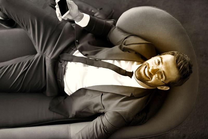 Male wearing eyeglasses sitting on sofa looking up at the camera with smartphone in right hand