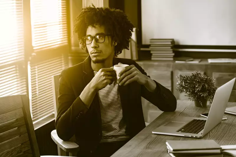 Male wearing eyeglasses in an office holding a coffee cup looking out of window