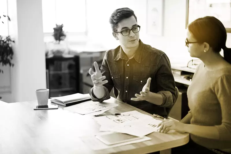 Male wearing eyeglasses speaking with his hands to a female wearing eyeglasses at a table