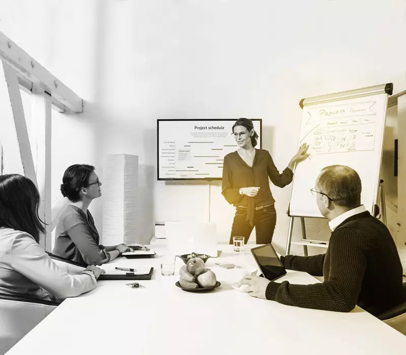 Female wearing eyeglasses pointing at a board conducting a meeting with three other people