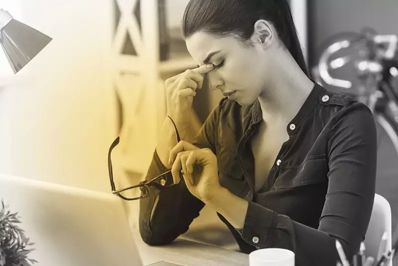 Female holding glasses in left hand pinching her forehead with right hand