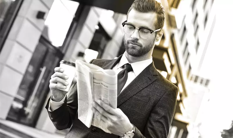 Male wearing glasses holding a coffee and reading a newspaper