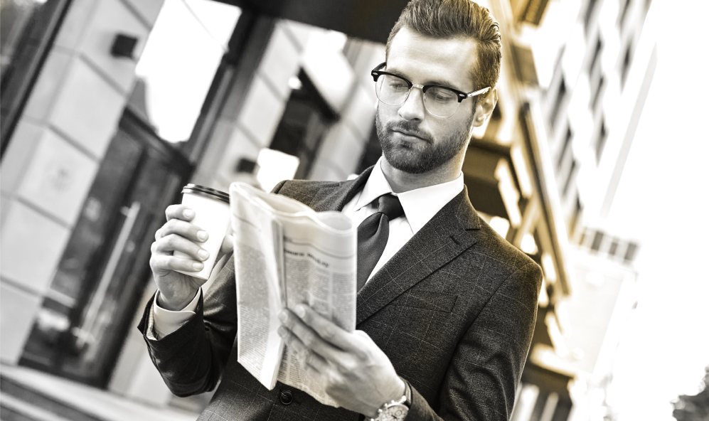 Male wearing glasses holding a coffee and reading a newspaper