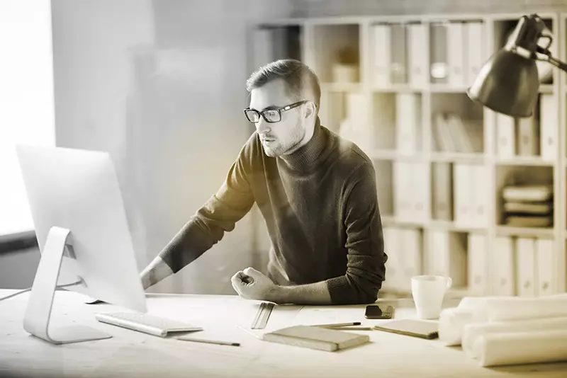 Male working indoors wearing eyeglasses behind computer screen