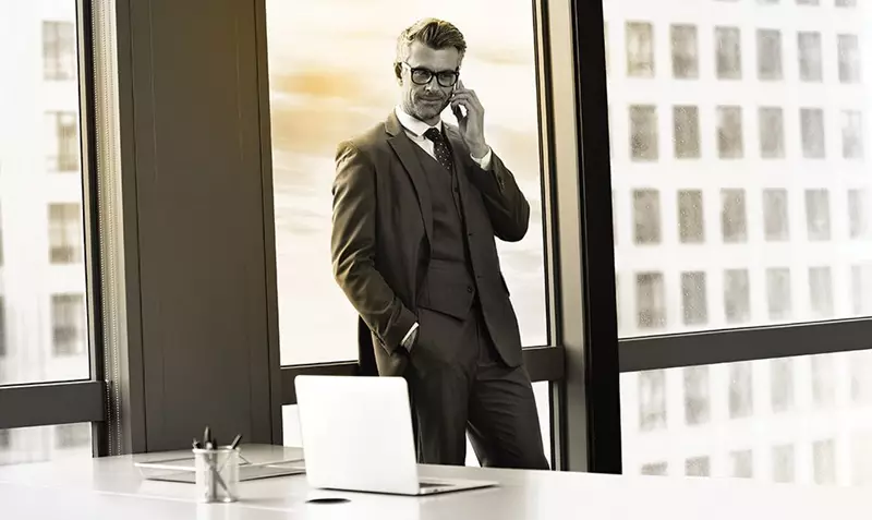 Business man in suit wearing glasses standing behind desk on a phone call